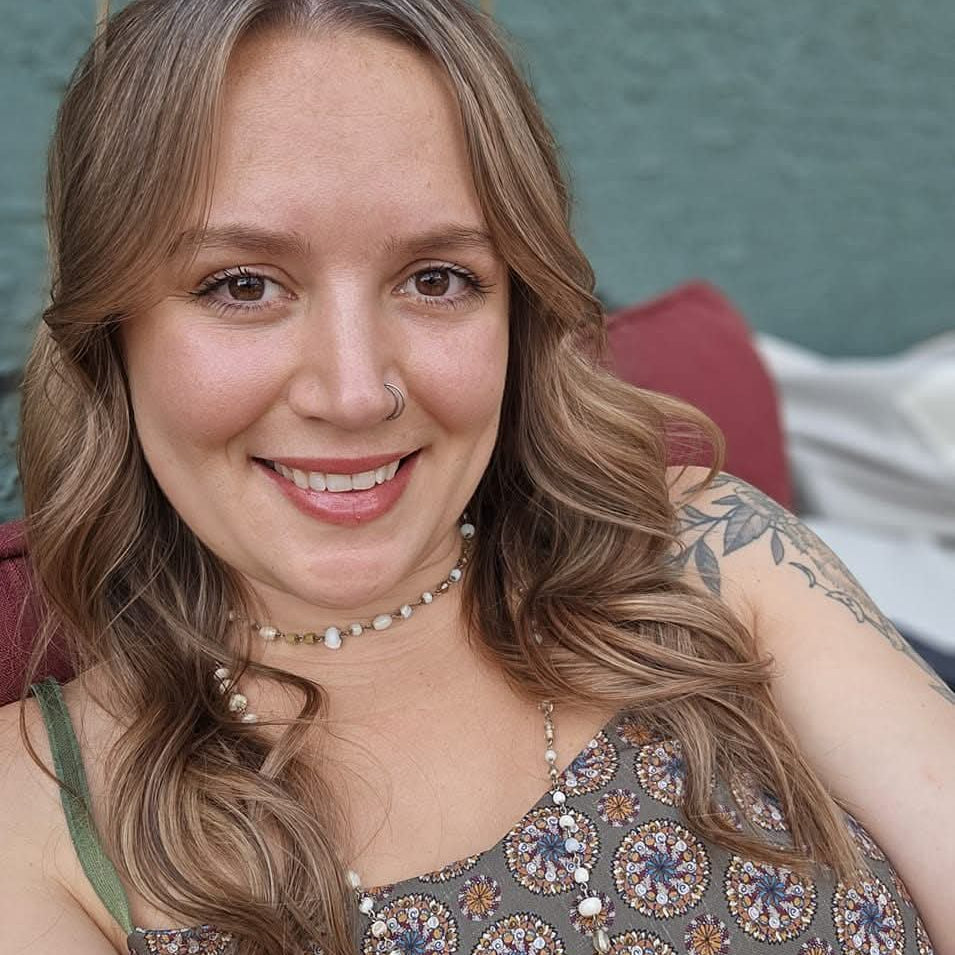 Portrait of the artist behind Moonlit Moss Studio smiling outdoors with wavy hair, floral tattoos, and earthy bohemian clothing.
