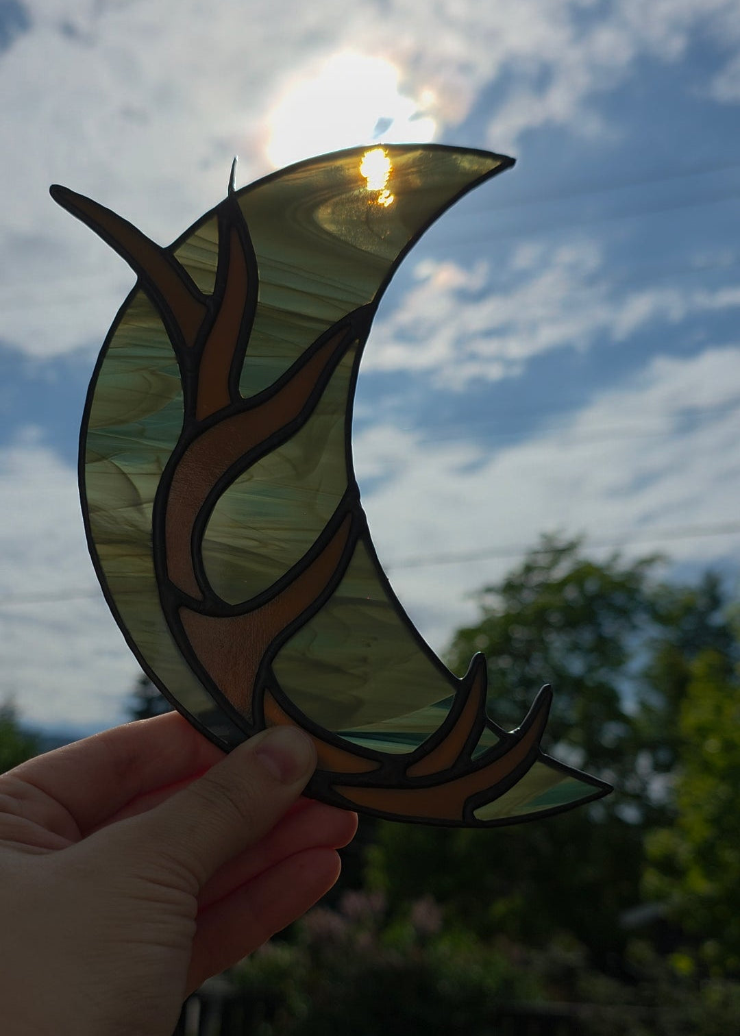 A hand holds a stained glass crescent moon ornament with brown antler-like designs, set against a bright outdoor sky with clouds and greenery.